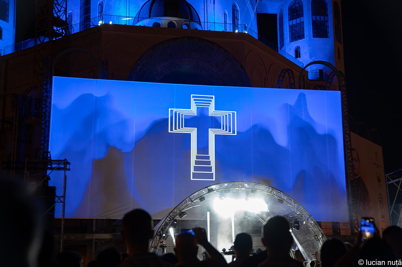 A cross projected on a large screen above the crowd at a Padre Guilherme cathedral square set