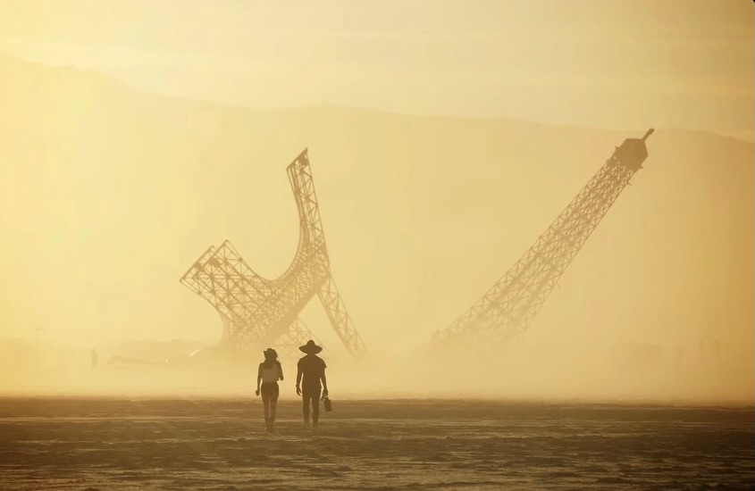 Burners walking toward the broken Eiffel Tower at Burning Man 2026 through desert dust